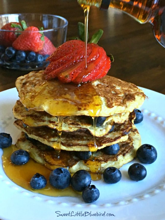 Photo of 4 banana pancakes stacked on top of each other topped with a sliced strawberry and fresh blueberries on a white plate with maple syrup being drizzled on top.