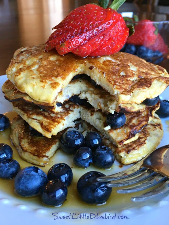 Photo of 4 banana pancakes served with a sliced strawberry and fresh blueberries with maple syrup on a white plate with a fork.