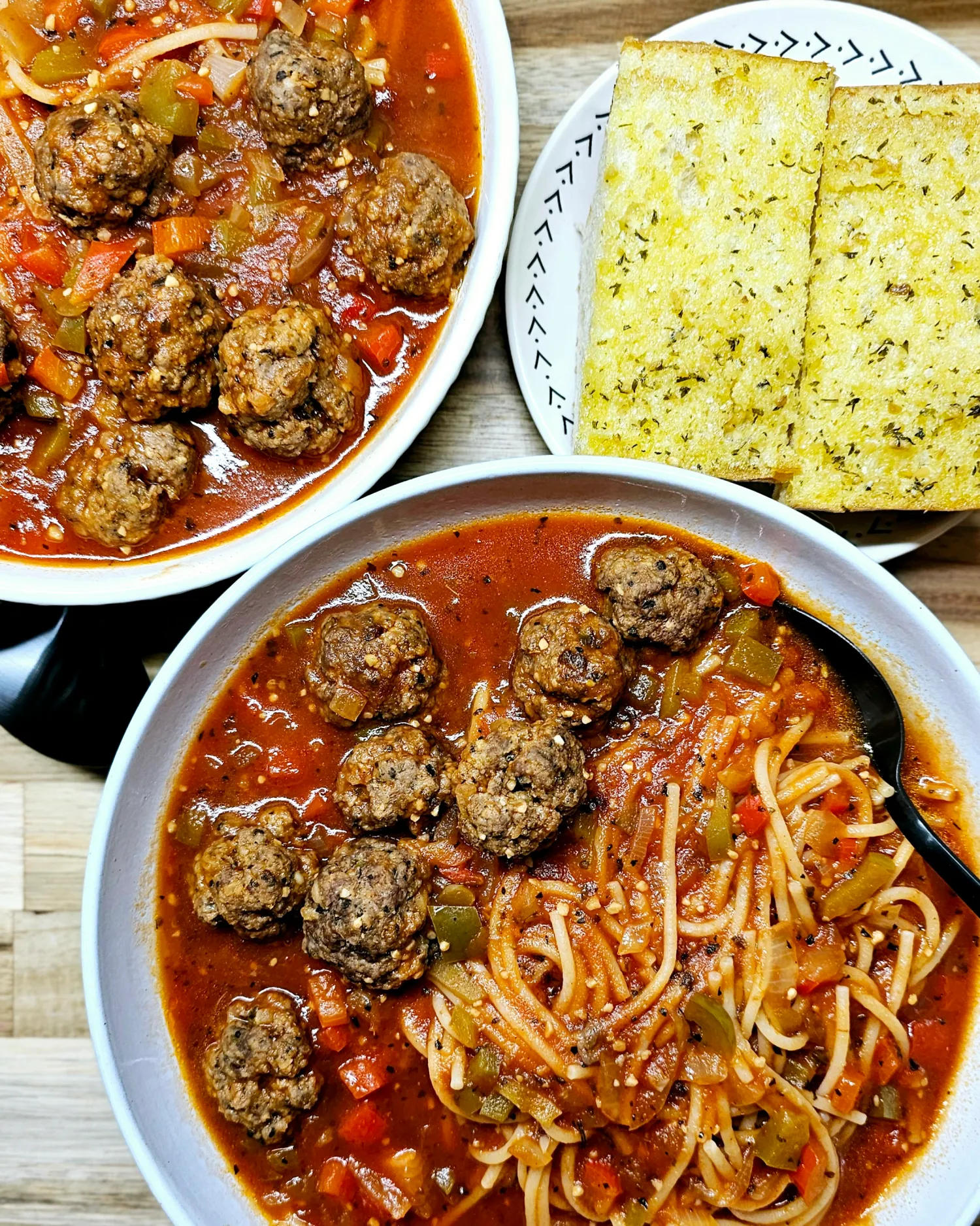 This photo shows two white bowls filled with spaghetti and meatball soup and a side of garlic bread on a plate.