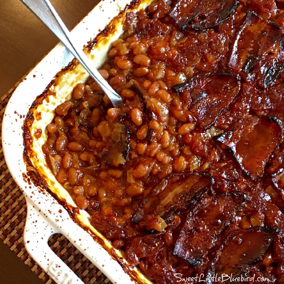This is a photo of the Best Baked Beans (0ven or Slow Cooker), after baking in a white casserole dish with a spoon, ready to serve. 