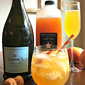 This photo shows a bottle of prosecco and bottle of apple cider. Next to them are two glasses of apple cider mimosa spritzers. One is served in a champagne glass, the other is served in a short wine glass with no stem.