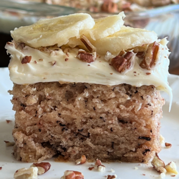 This is a photo of a square piece of Banana Bread Crazy Cake topped with vanilla frosting, chopped pecans and fresh sliced banana served on a white plate.