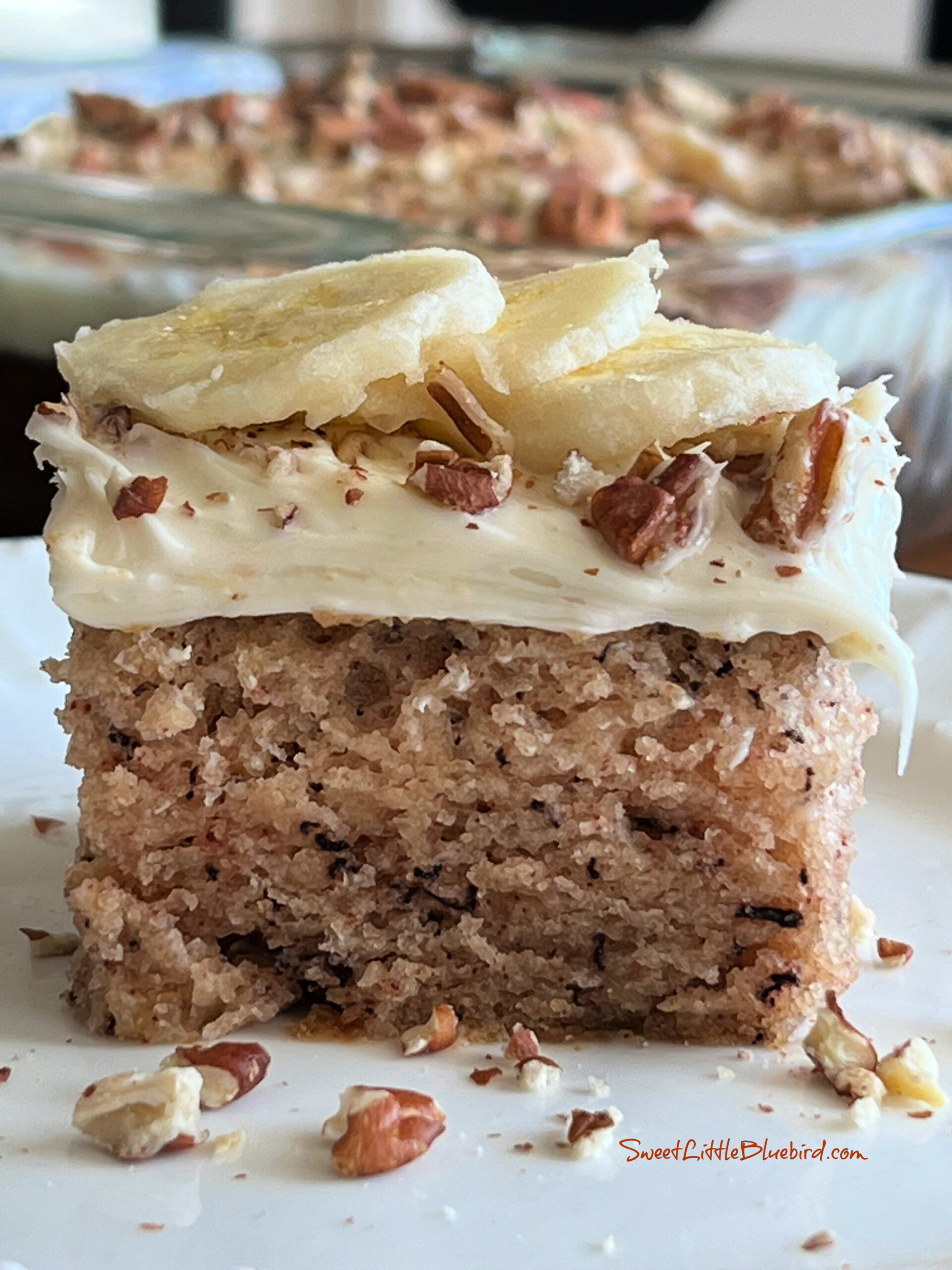 This is a photo of a square piece of Banana Bread Crazy Cake topped with vanilla frosting, chopped pecans and fresh sliced banana.