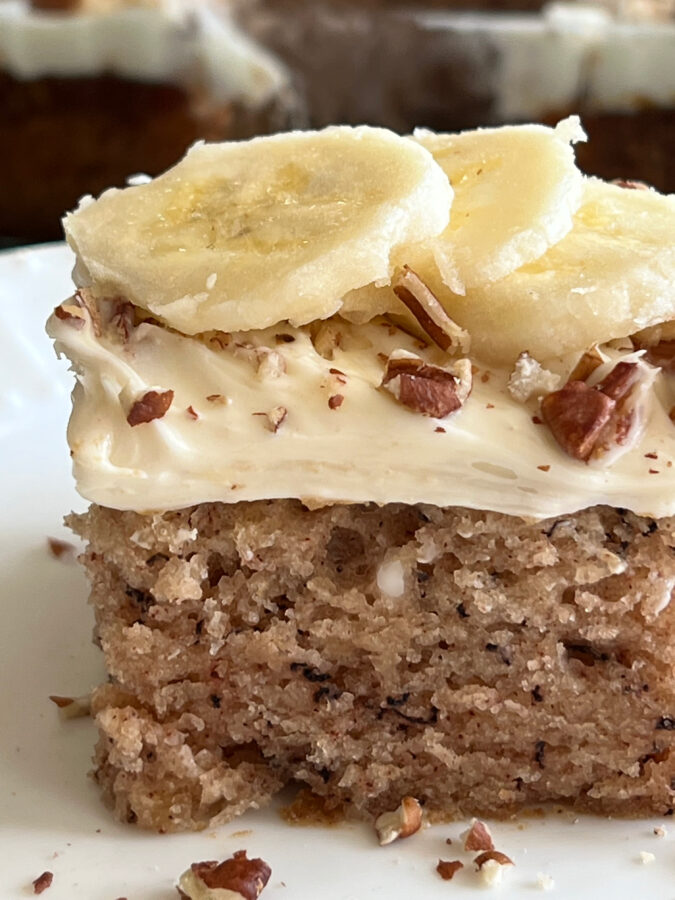 This is a photo of a square piece of Banana Bread Crazy Cake topped with vanilla frosting, chopped pecans and fresh sliced banana served on a white plate. 