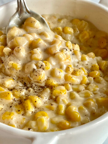 This is a photo of Homemade Creamed Corn served in a white serving dish with a spoon.
