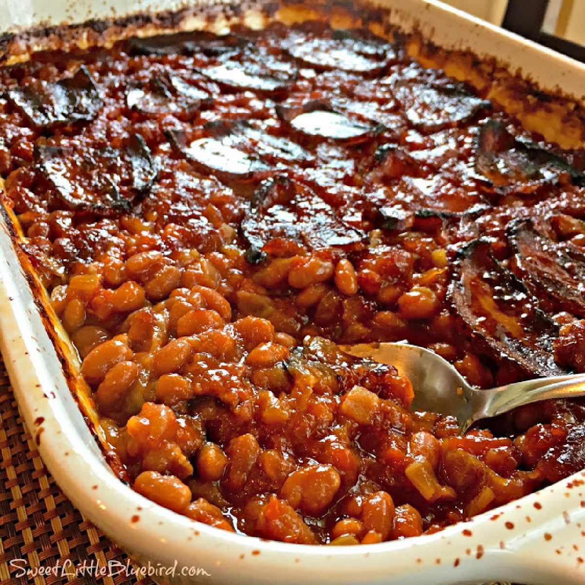 This is a photo of the Best Baked Beans (0ven or Slow Cooker), after baking in a white casserole dish with a spoon, ready to serve. 