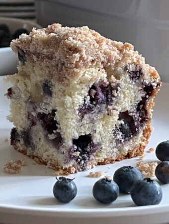 Piece of Blueberry Buckle Coffee Cake served on a white plate with 6 fresh blueberries scattered on the plate.