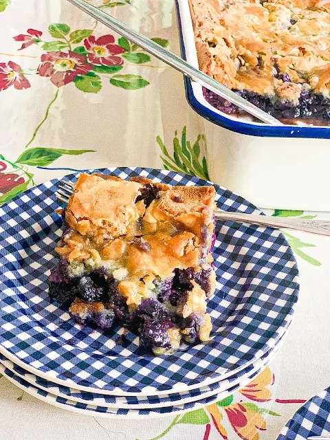 This photo shows Blueberry Gooey Butter Cake served on a plate with the baking dish behind it. 