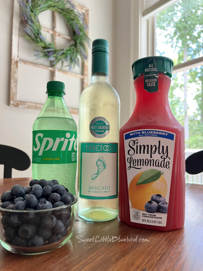 Photo of ingredients to make a Moscato Wine Spritzer - bottle of sprite, bottle of Moscato, blueberry lemonade and a small bowl of fresh blueberries.