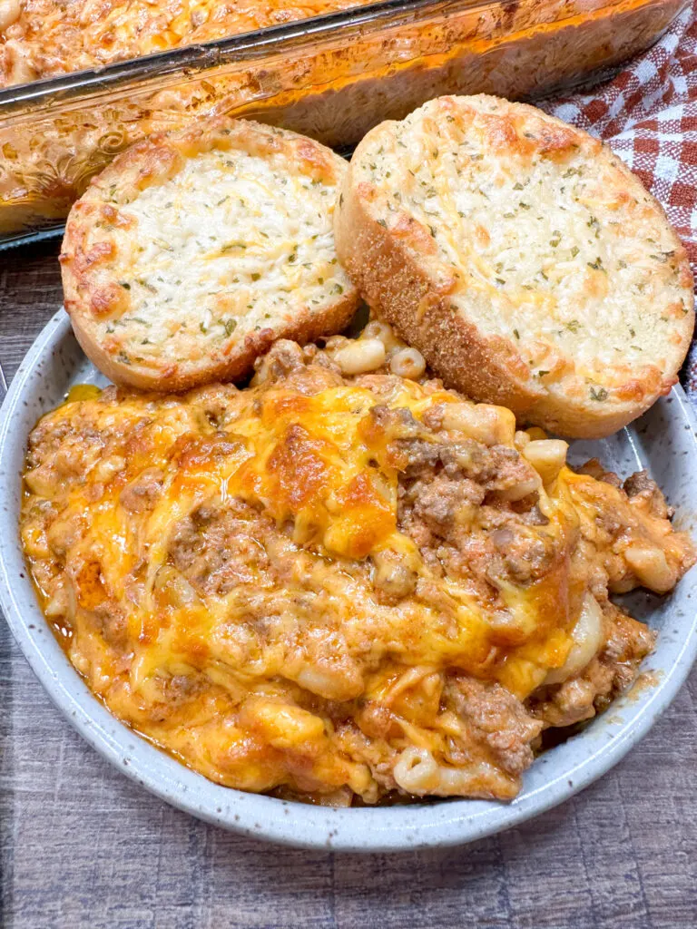 This photo shows hamburger casserole served in a bowl with cheese garlic bread on the side. 