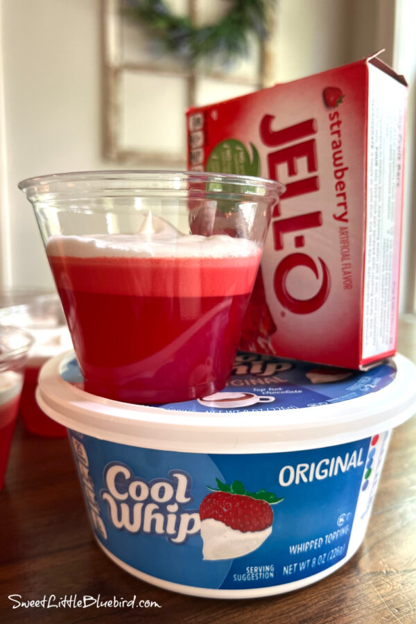 A photo of the jello dessert in a clear cup on top of a tub of Cool Whip next to a box of Strawberry Jello.