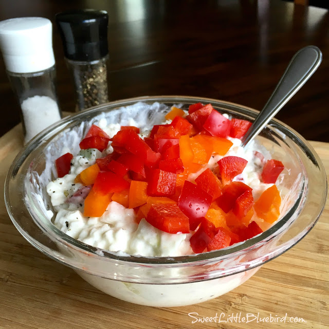 This photo shows chopped red pepper on top of cottage cheese in a clear glass mixing bowl with a spoon. 