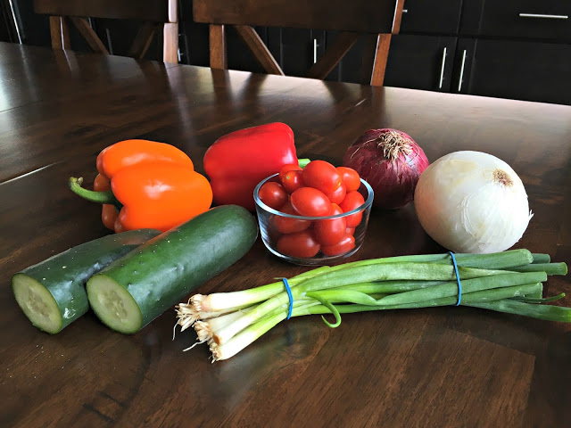 This photo shows veggies on a kitchen table - An orange and red pepper, cucumber. Cherry tomatoes and red and white onion.