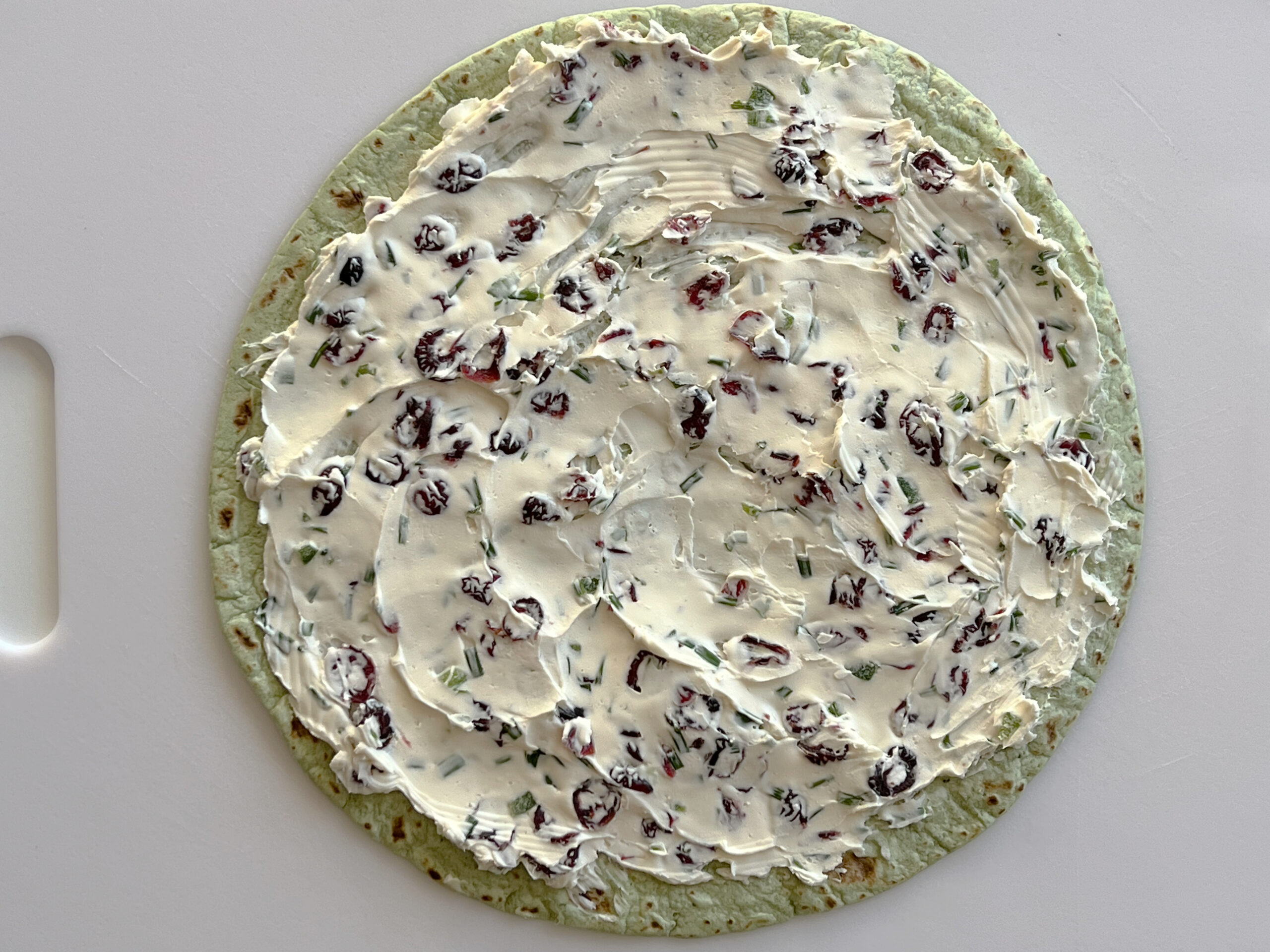 This photo shows the cream cheese mixture spread evenly on a spinach herb tortilla on a white cutting board.  