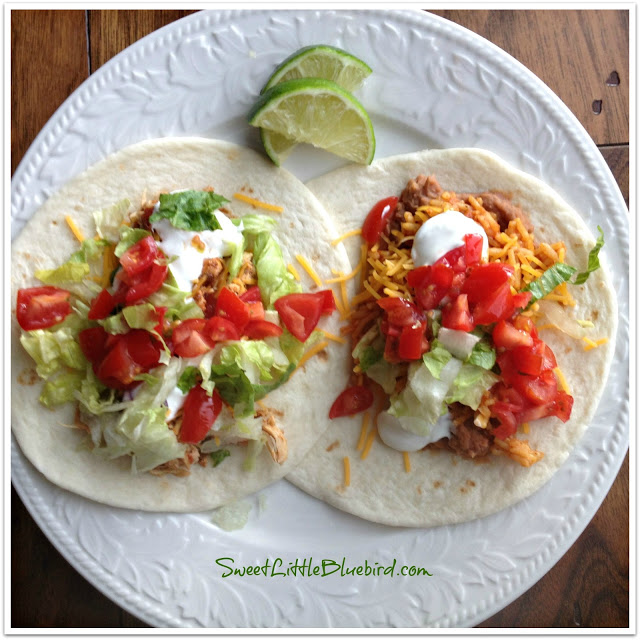 This photo shows 2 soft tacos on a white plate, topped with toppings,- diced, tomato, chopped lettuce, shredded cheddar cheese, sour cream, with 2 sliced limes, ready to eat. 