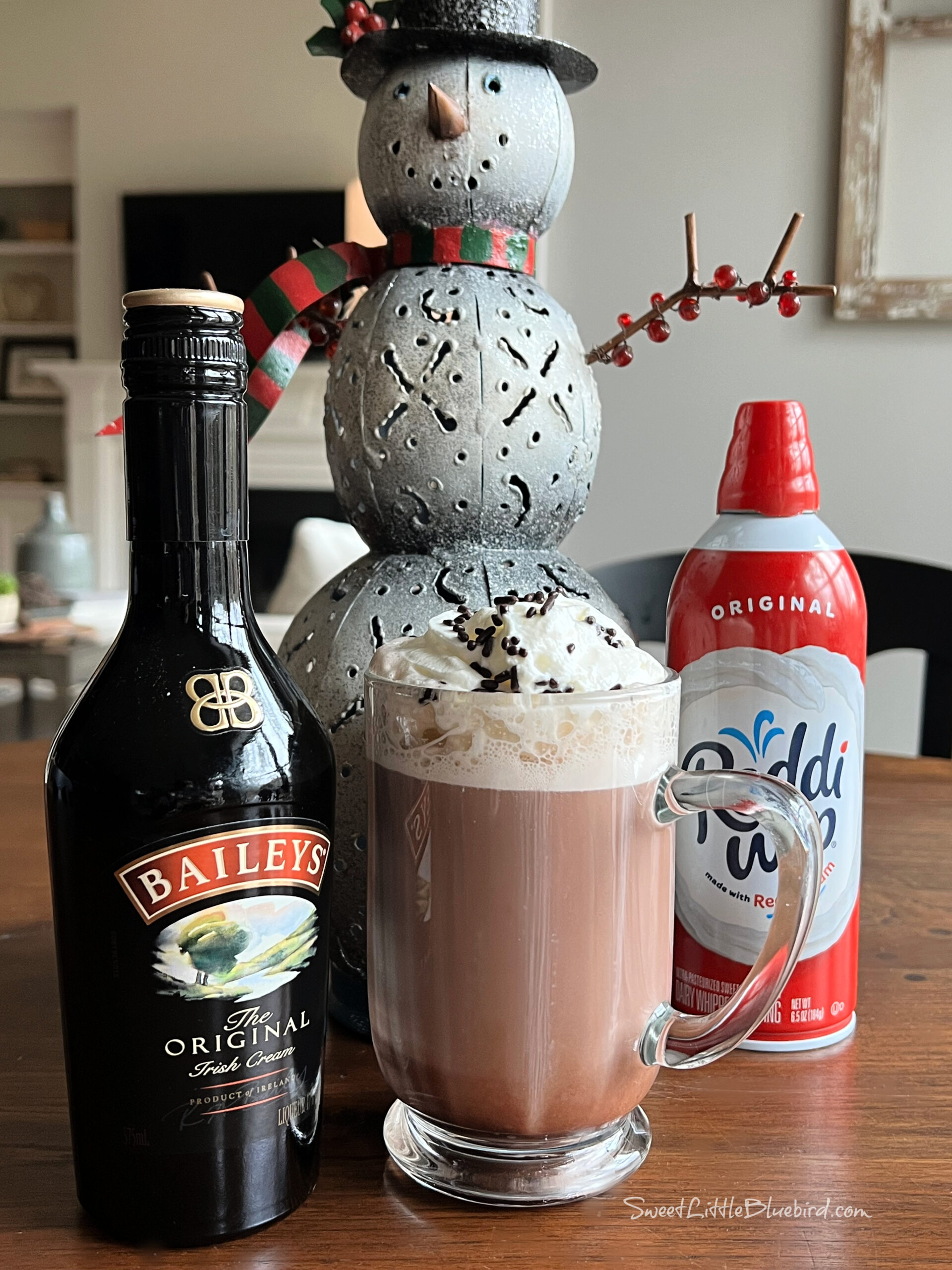 This photo shows a Dirty Snowman cocktail served in a clear mug glass, topped with whipped cream and chocolate sprinkles. Behind the mug is a decorative snowman figurine. Nest to the mug is a bottle of Baileys and a can of whipped cream.