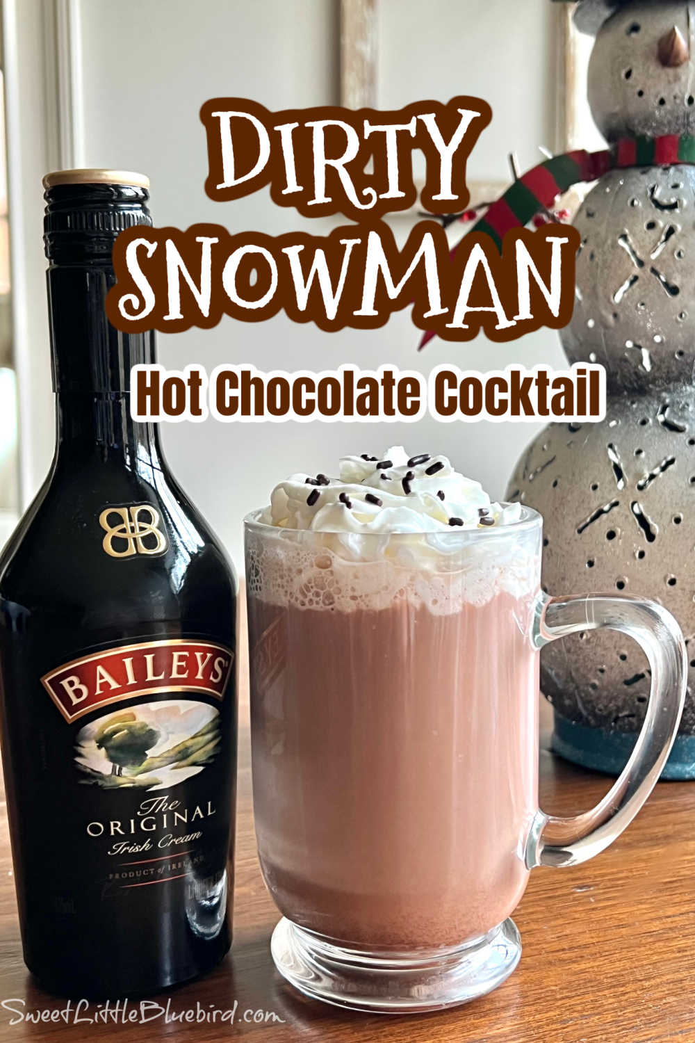 This photo shows a Dirty Snowman cocktail served in a clear mug glass, topped with whipped cream and chocolate sprinkles. Next to the mug is a bottle of Baileys Irish Crème.