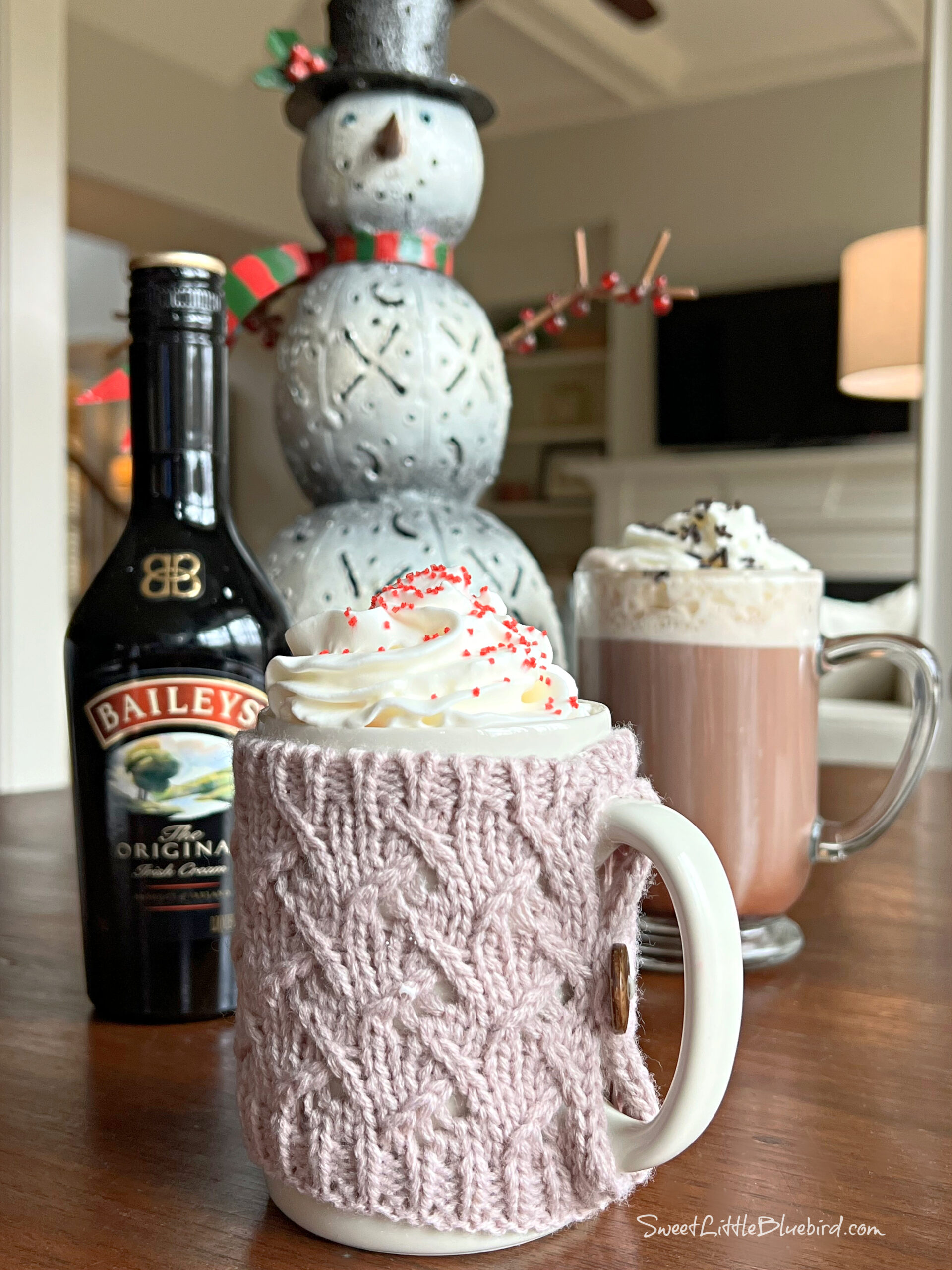 This photo shows the drink served in a mug with whipped cream and red sprinkles. Behind the mug is another drink in a clear glass mug next to a snowman figurine and a bottle of Baileys Irish Crème. 