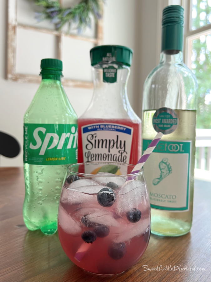 Photo of a moscato wine spritzer in a short wine glass made with Blueberry Lemonade garnished with fresh blueberries with a purple and white straw. A bottle of sprite, blueberry lemonade and moscato are behind the glass.