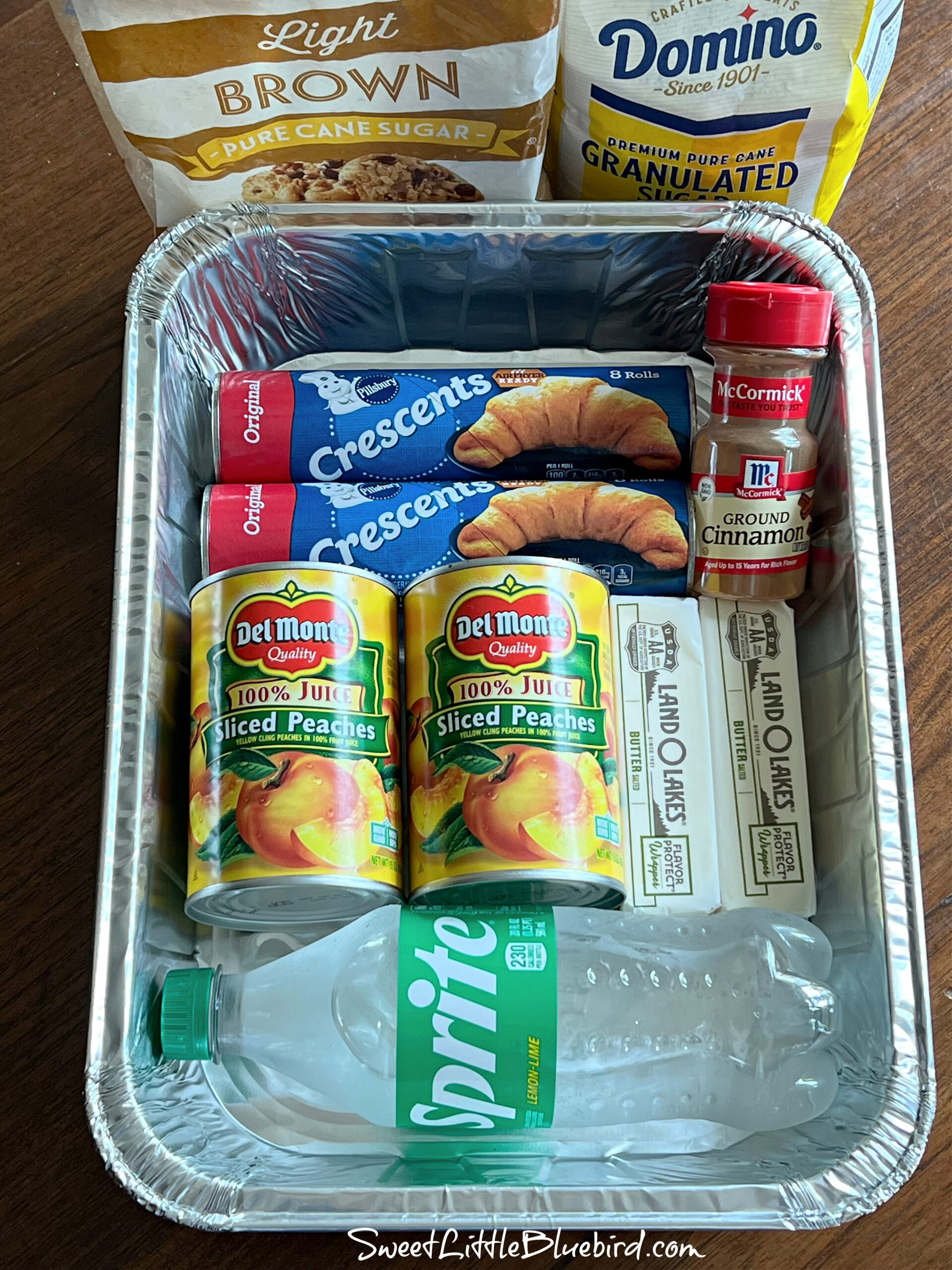 This photo shows the ingredients needed to make the dumplings inside a disposable 9x13 baking aluminum pan (cans of peaches, crescent rolls, butter, cinnamon and a bottle of Sprite). Next to the pan is a bag of granulated sugar and a bag of brown sugar.
