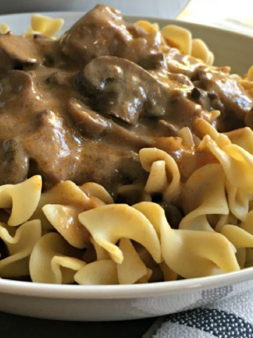 This photo shows beef stroganoff served over egg noodle in a white bowl.