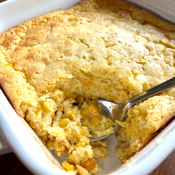 This is a photo of Easy Corn Casserole baked in a white square baking dish with a spoon ready to serve.