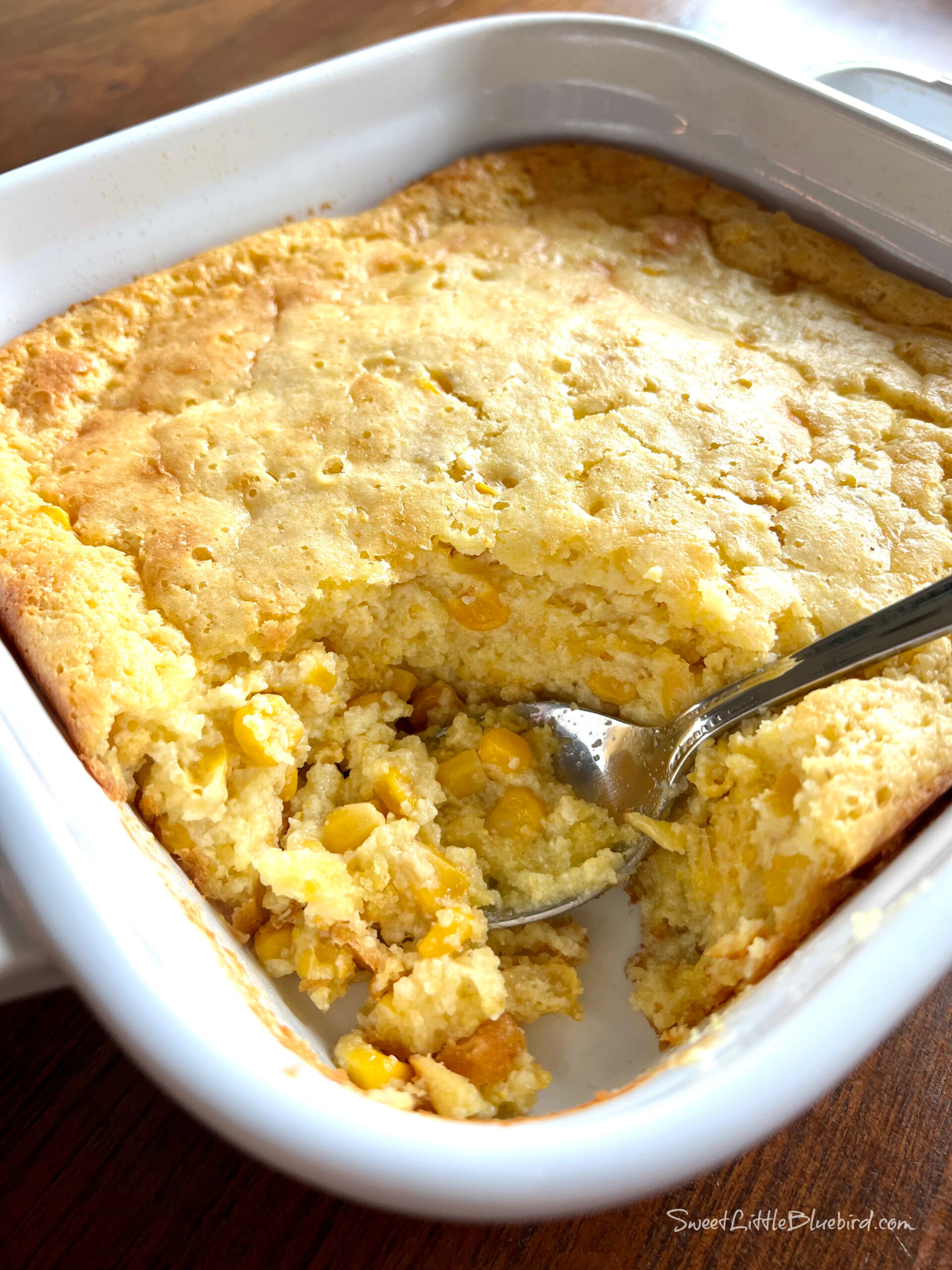 This is a photo of Jiffy Corn Casserole baked in a square white baking dish with a spoon ready to serve.