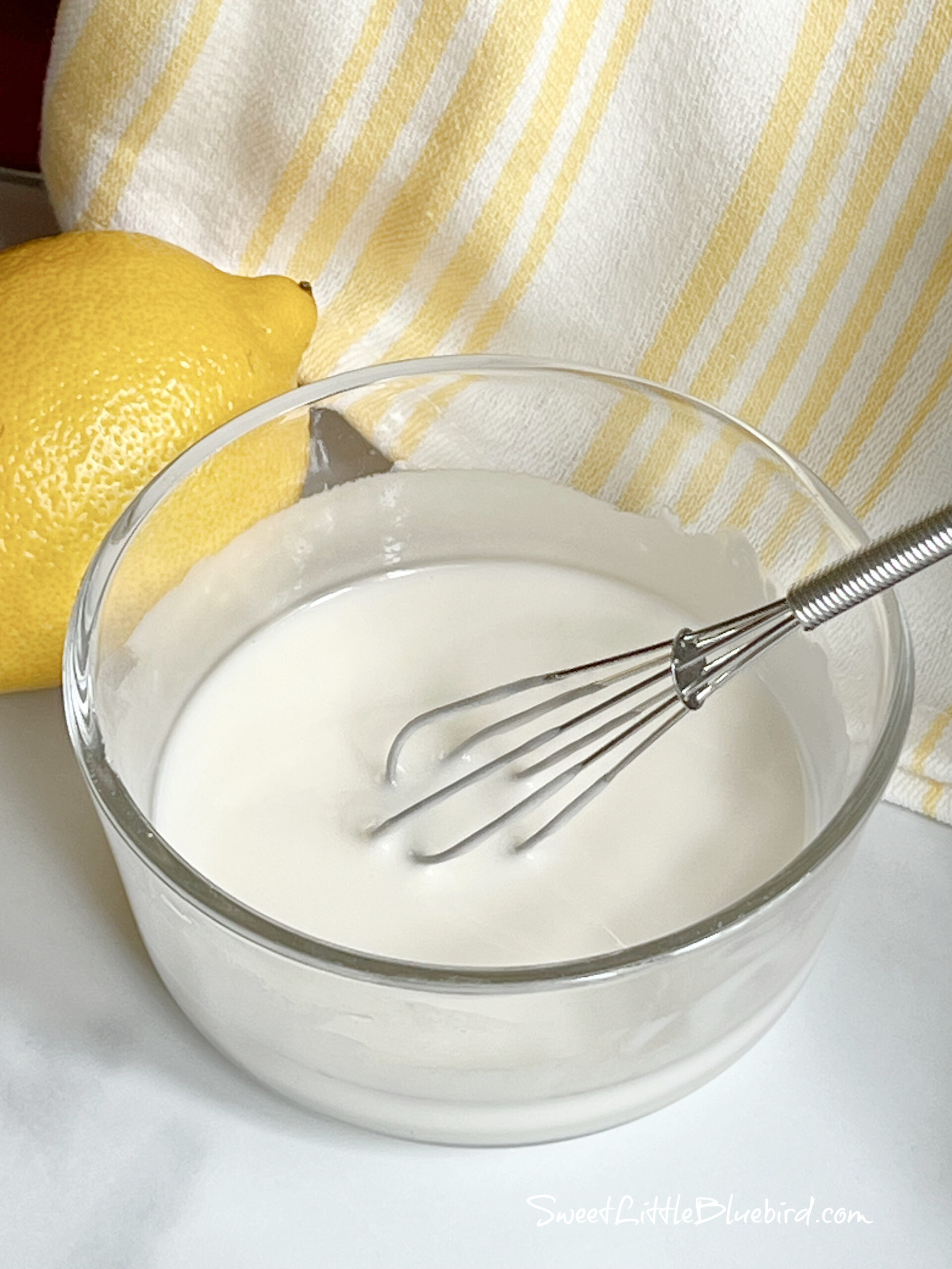This photo shows lemon glaze in a small clear glass bowl with a small whisk.