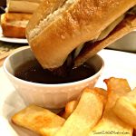 This image shows a French dip sandwich dipped in a small bowl of au jus (broth). The sandwiched is served with steak fries.