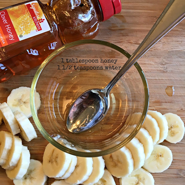 This photo shows a small bowl with a mixture of honey and water with a spoon, and a bottle of honey and sliced bananas next to the bowl.