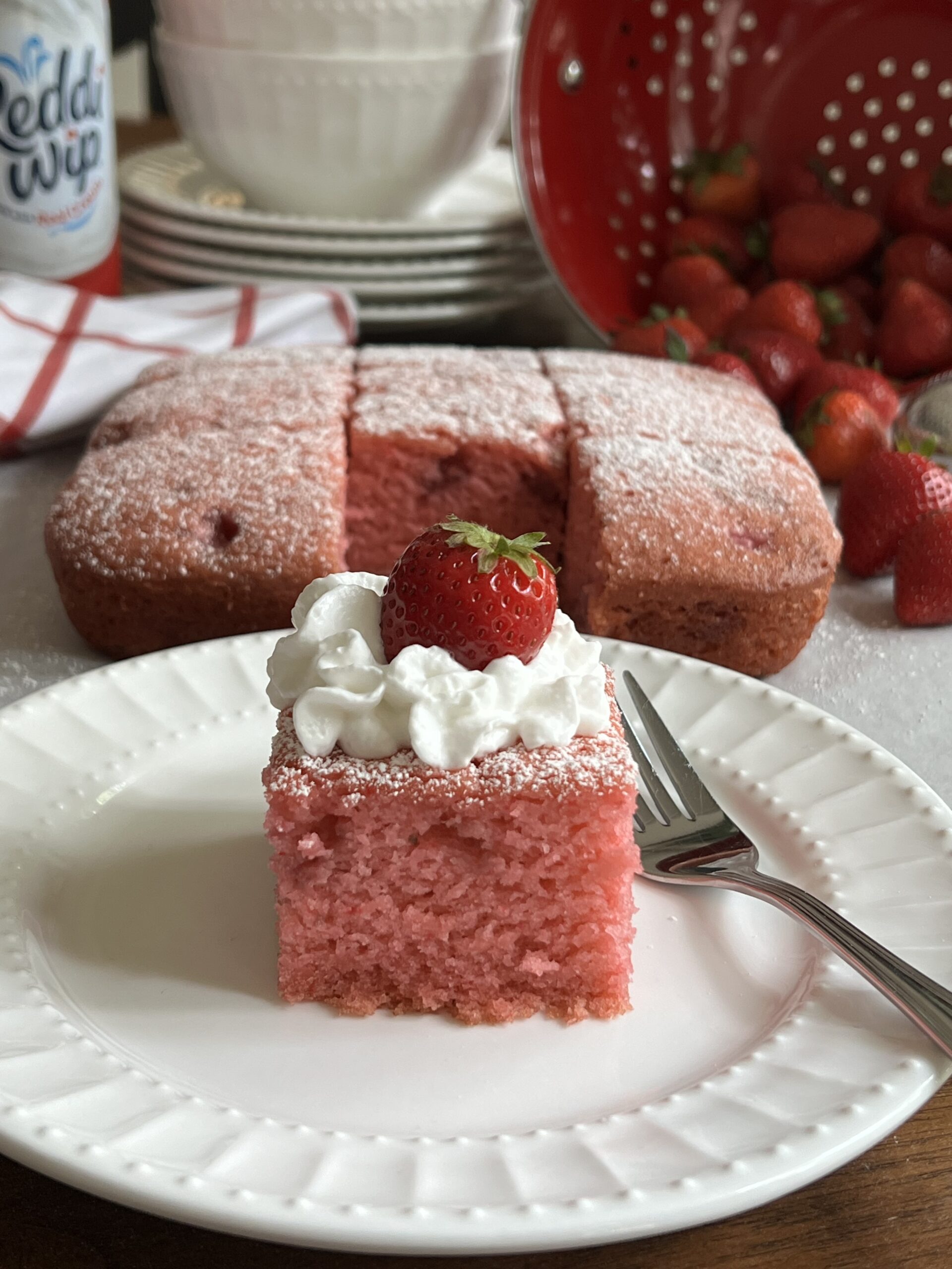 Strawberry Crazy Cake on a white plate with a dusting of powdered sugar, a dollop of whipped cream and a fresh strawberry. Behind the plate is the cake, sliced with fresh strawberries in a red colander, more plates and bowls.