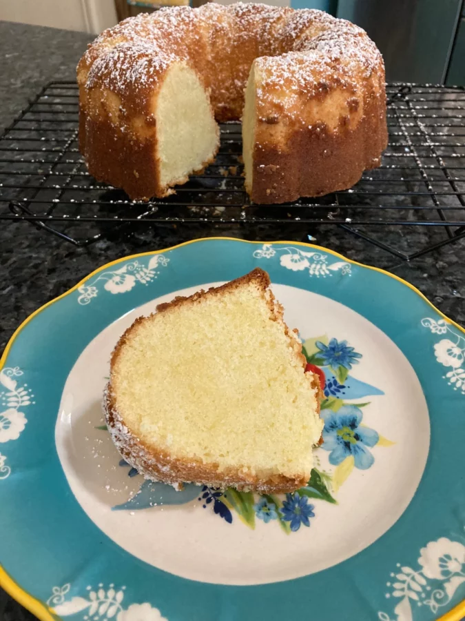 Photo of a slice of pound cake on a plate with the cake shown behind on a cooling rack - by Out of the Box Baking