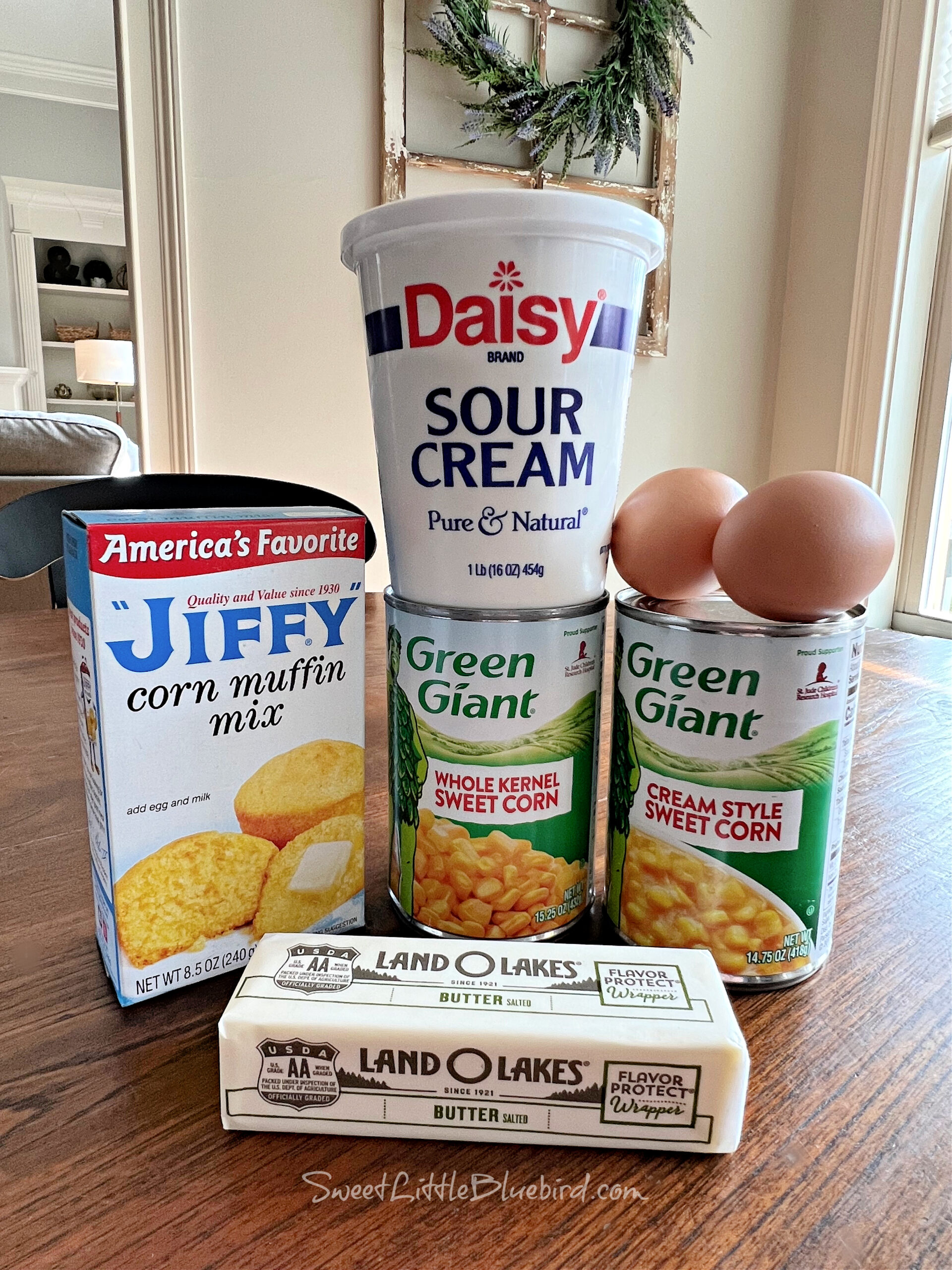 This is a photo of the ingredients needed to make the corn casserole placed neatly together on a table - a box of Jiffy Corn Muffin Mix, container of sour cream, stick of butter, 2 eggs, can of cream style corn and a can of whole kernel corn. 