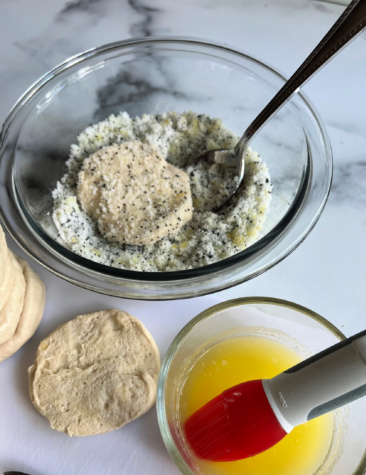 This photo shows a small bowl of melted butter with a brush, a biscuit next to it and a biscuit being coated in the sugar lemon poppy seed mixture in a small clear glass mixing bowl. 