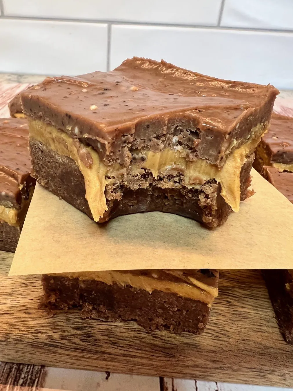This is a photo of Layered Peanut Butter Brownies on a brown cutting board, cut into squares with two stacked on top of each other. The top brownie has a bite missing. Photo by The House on Silverado. 