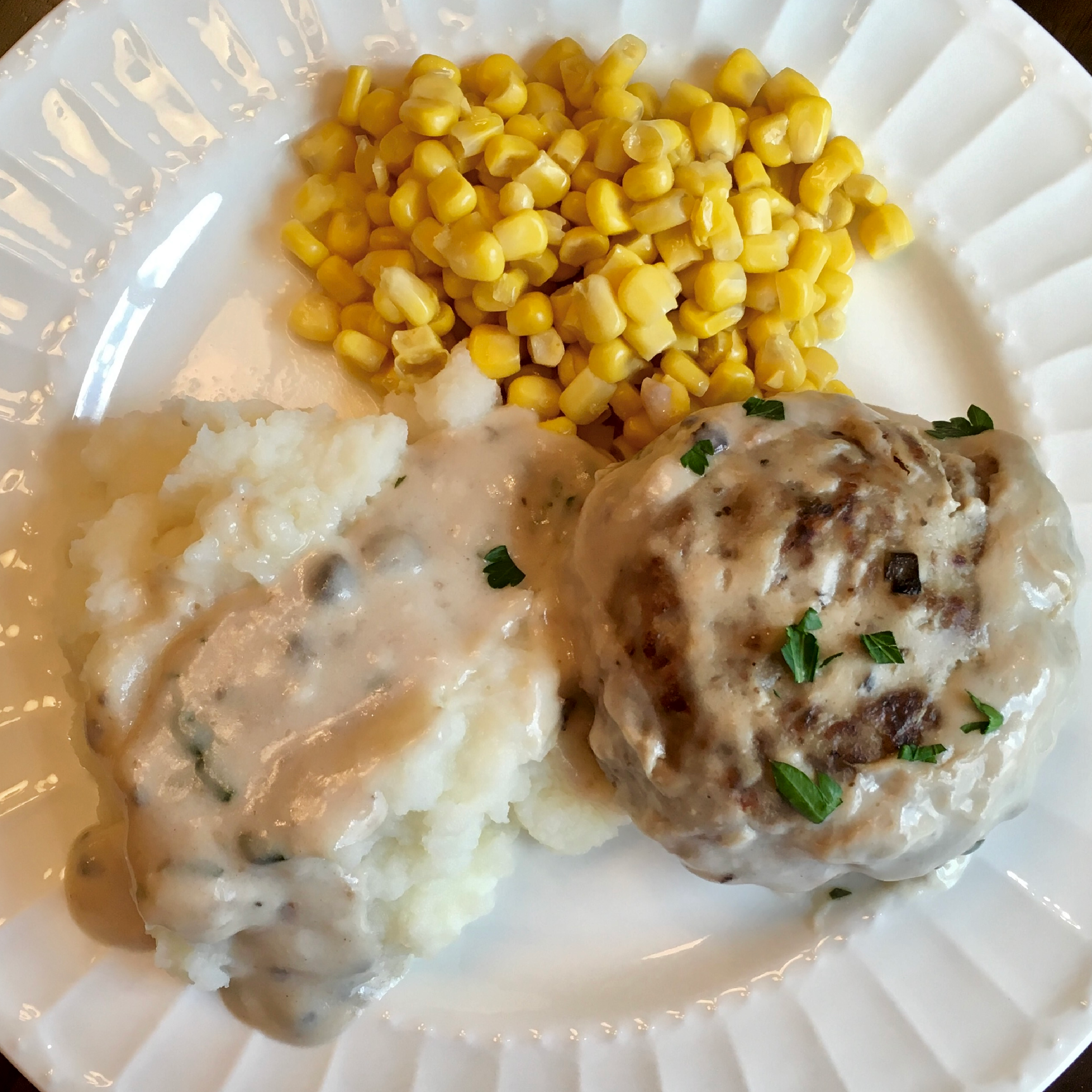 This is a photo of a plate served with a hamburger steak served with mashed potatoes and corn on a white plate. 