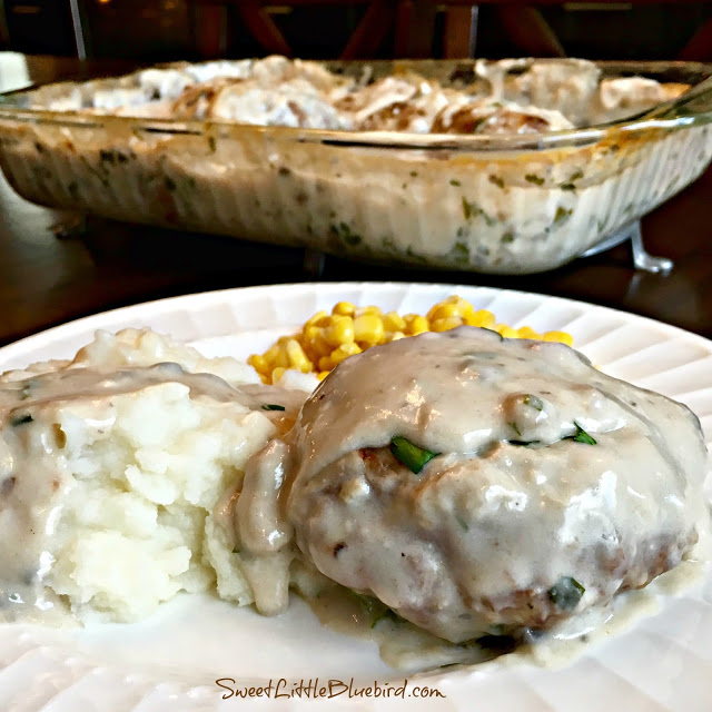 This is a photo of a Hamburger Steak served on a white plate next to mashed potatoes and corn. The steak and mashed potatoes are topped with gravy. Behind the plate is the baking pan with the hamburger steaks.
