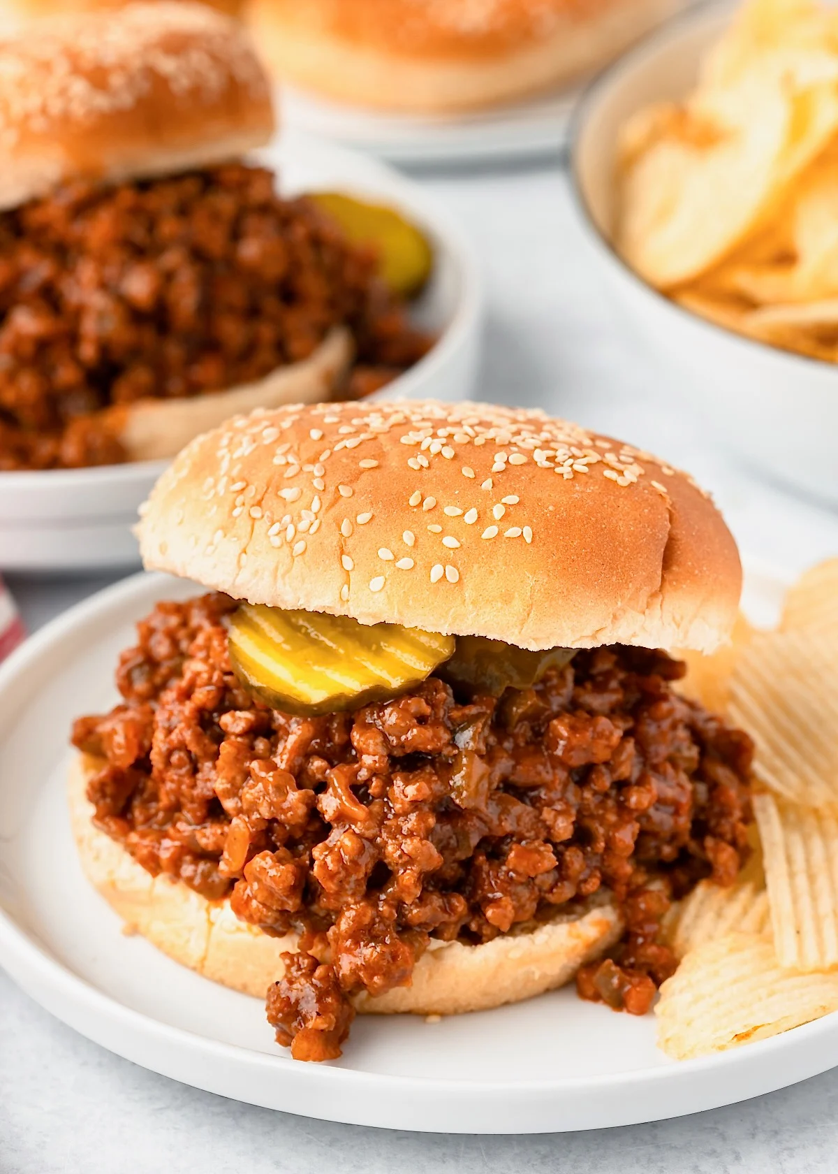 This photo shows a Sloppy Joe sandwich on sesame bun with pickles on a white plate with potato chips. 