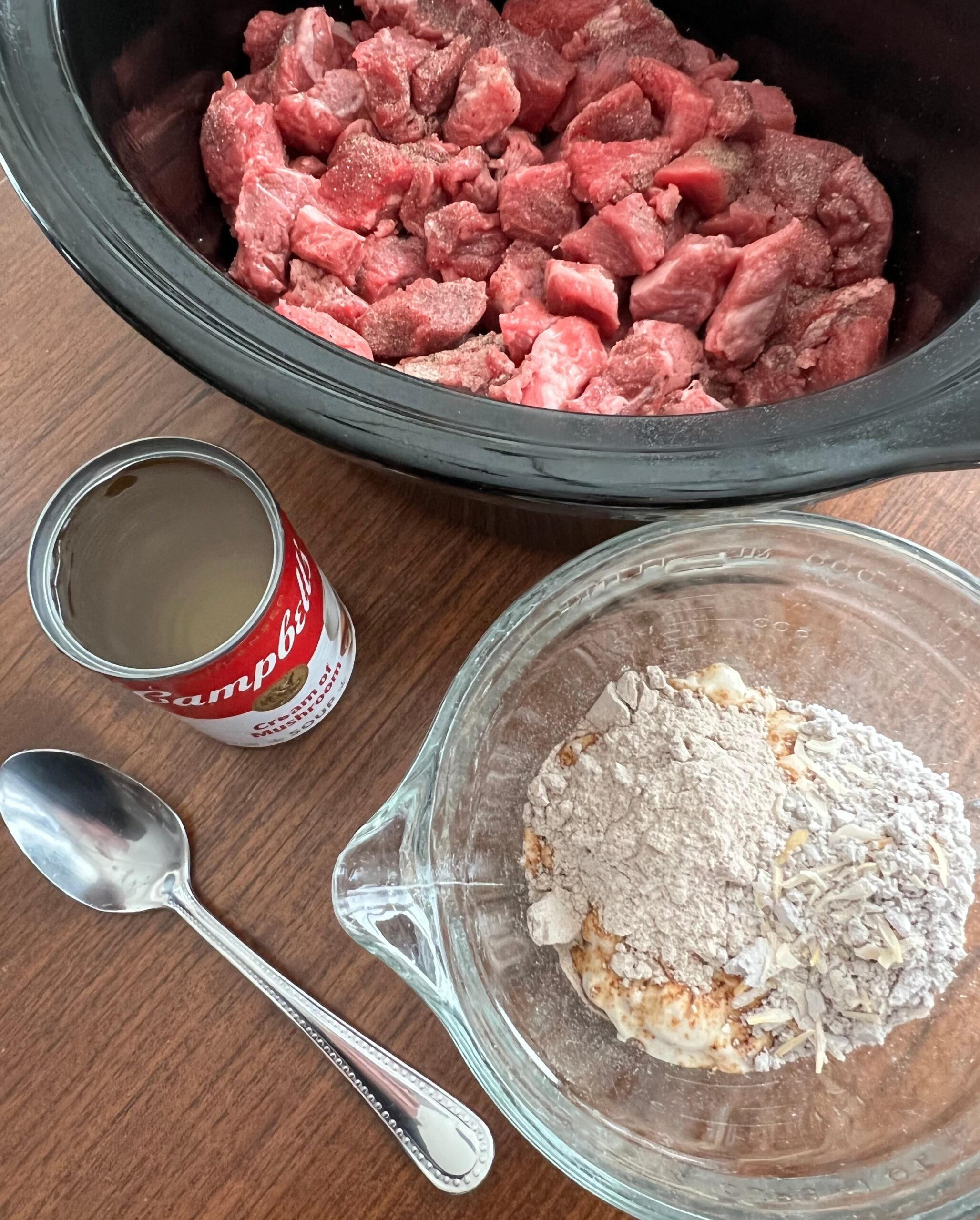 This picture shows the Beef Tips being made in a slow cooker. This is a photo of a slow cooker dish filled with the stew beef with a measuring bowl next to it, filled with the wet and dry soups and the dry gravy mix. Next to them is the soup can filled with water with a spoon next to the bowl.