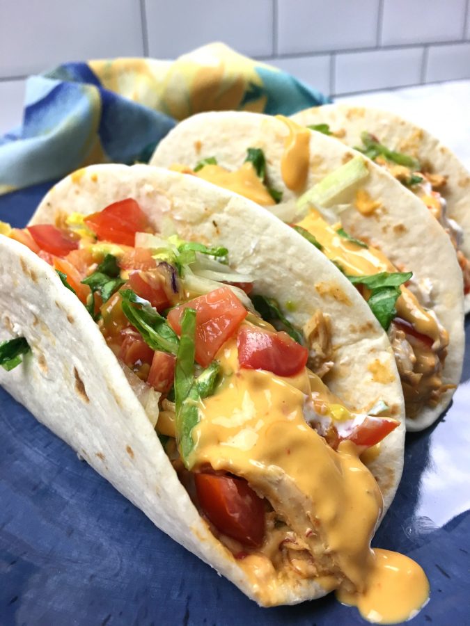 This is a photo of 3 queso chicken tacos served on a blue plate in soft taco flour shells topped with lettuce and tomato and extra queso. 
