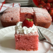 Strawberry Crazy Cake on a white plate with a dusting of powdered sugar, a dollop of whipped cream and a fresh strawberry. Behind the plate is the cake, sliced with fresh strawberries in a red colander, more plates and bowls.