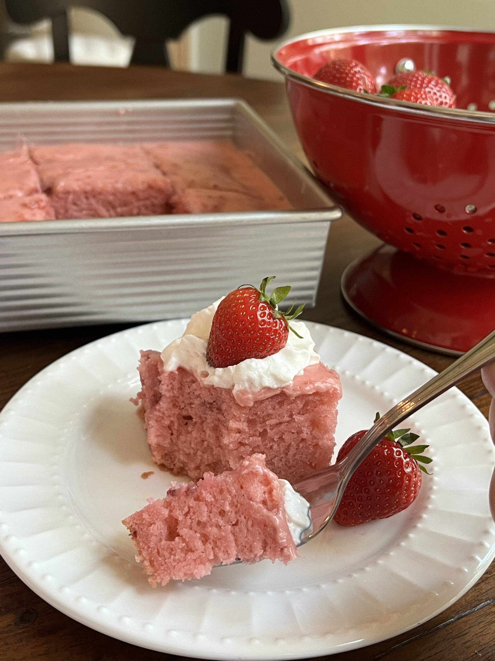 A piece of strawberry cake on a white plate topped with strawberry glaze, whipped cream and a fresh strawberry on top and one on the side. Behind the cake is a red colander and the cake in the baking pan.
