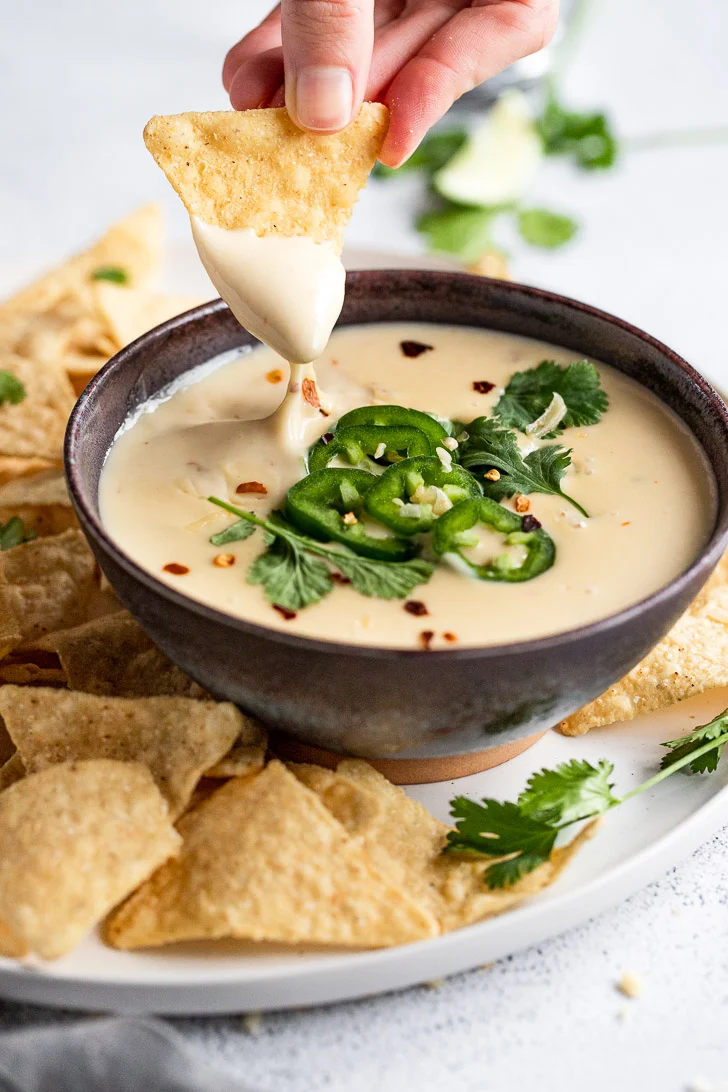 This is a photo of queso served in a bowl on top of a plate with tortilla chips, with a hand dipping a chip in the queso.