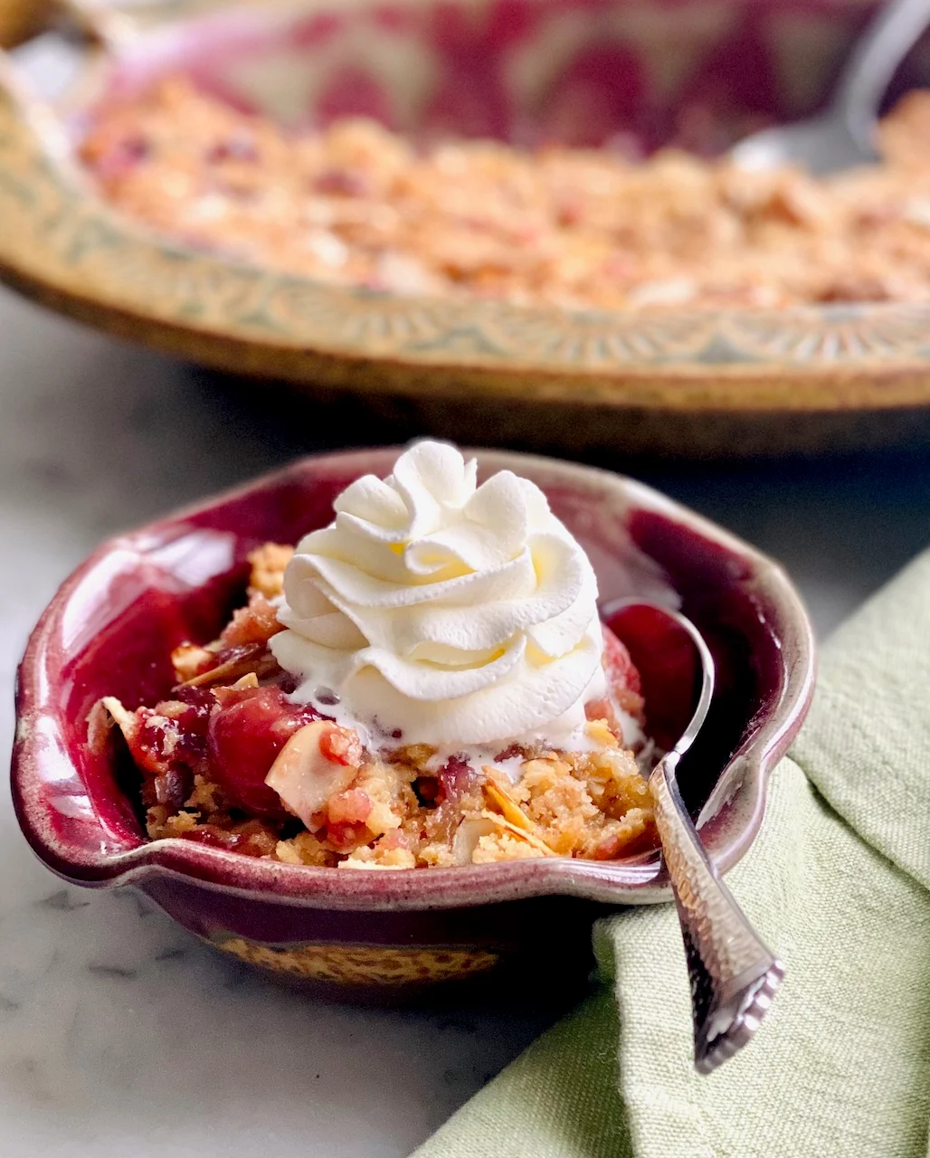 This is a photo of Cherry Almond Cobbler served in a red bowl with a spoon topped with a scoop of vanilla ice cream. Behind the bowl is the cobbler in a pie plate.