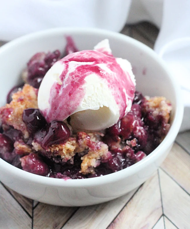 This is image shows Cake Mix Fruit Cobbler served in a white bowl topped with a scoop of vanilla ice cream. 