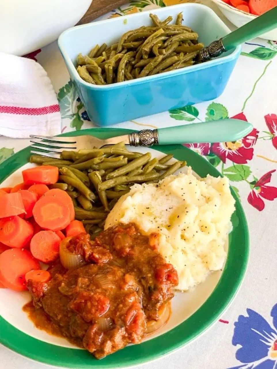 Swiss steak served on a plate with a side of mashed potatoes, green beans and carrots. Behind the plate is a blue serving dish with green beans. 