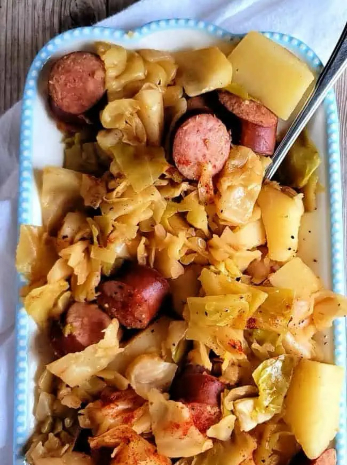 Cooked and ready to serve, crockpot Kielbasa with cabbage and potatoes in a blue and white rectangular serving dish with a spoon. 