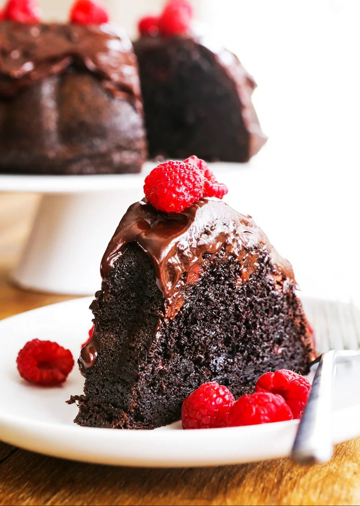 Chocoalte Rpasberry Bundt Cake A piece of Chocolate Raspberry Bundt Cake on a white plate. The cake is topped with two raspberries and there are several raspberries on the plate. Behind the plate is a white cake platter with the cake.