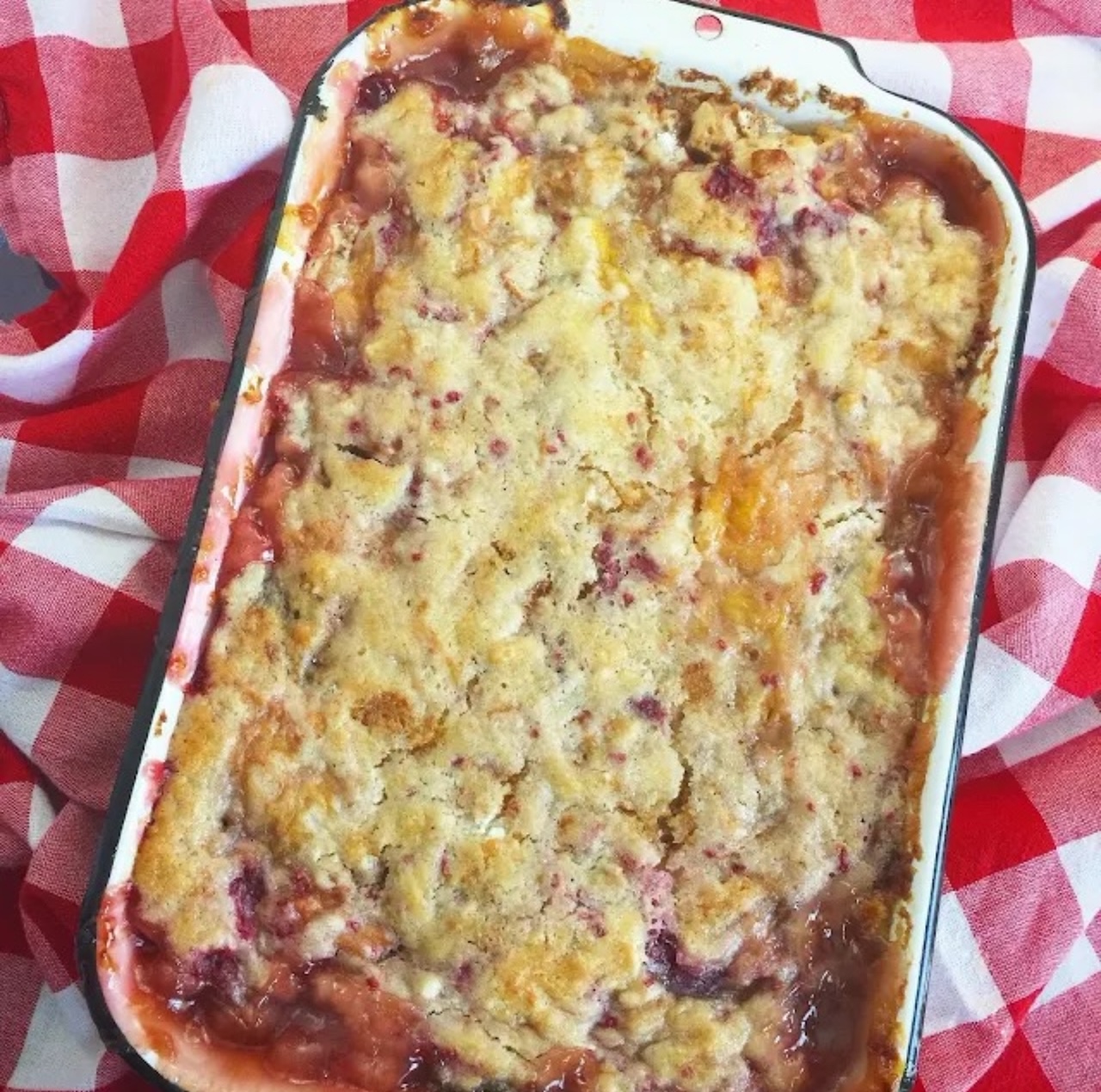 Sugar Cookie Peach Cobbler Sugar Cookie Peach Cobbler baked in a white baking dish placed on top of a red and white checkered table cloth, ready to serve.