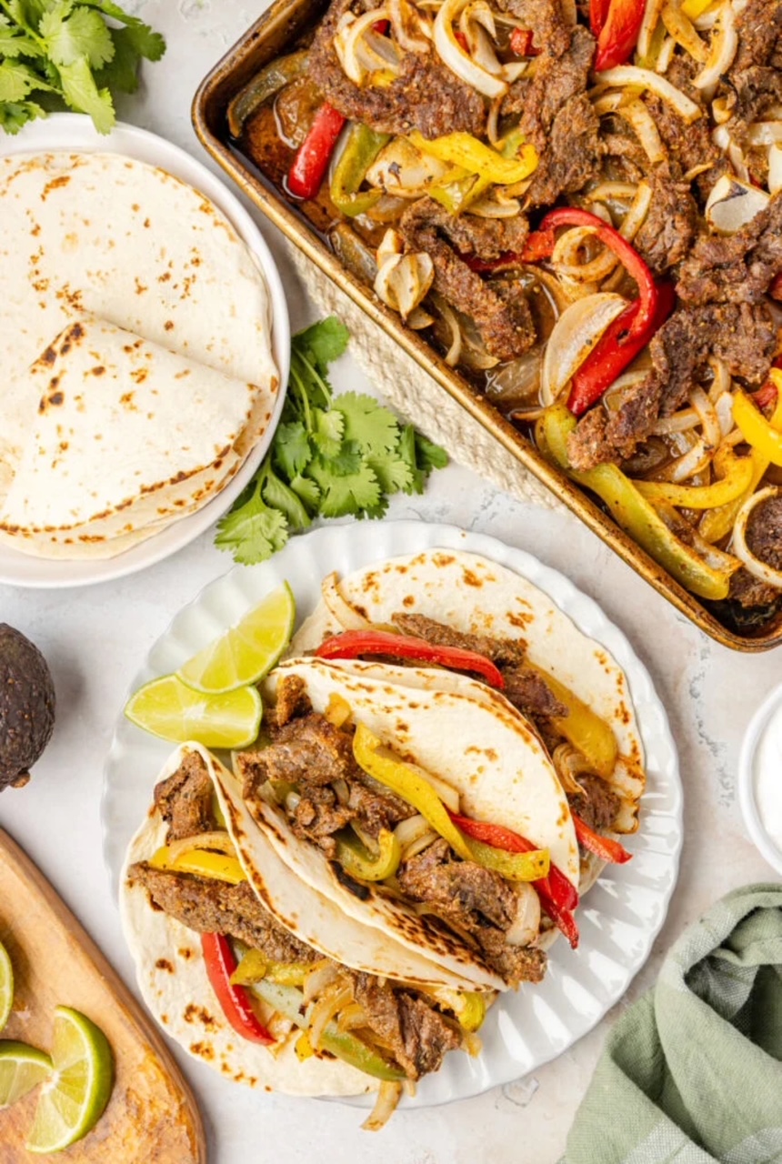 Sheet Pan Steak Fajitas Three Streak Fajitas on a white plate with two lime wedges. The plate is next to a sheet pan filled with the steak, peppers and onion. There is also a white plate with flour tortillas.